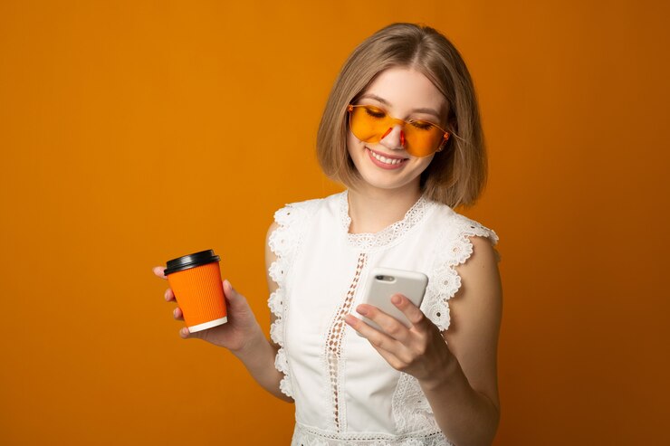 Mujer joven positiva en vasos amarillos con bebida y teléfono móvil