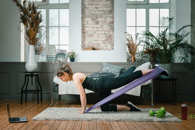 Mujer joven practica deportes en casa, entrenando en línea. La atleta balancea su pierna con gomas elásticas deportivas, mira una película y estudia desde una computadora portátil en la cama de fondo, florero, alfombra