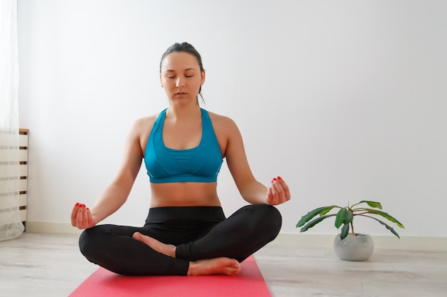 Mujer joven practica yoga en casa, meditando y sentado en una estera. Contra la pared blanca.