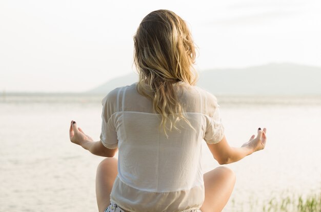 Mujer joven practica yoga y medita en posición de loto en la playa, frente al agua. Gran concepto de meditación y relajación.