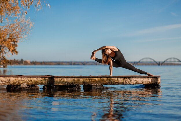 Mujer joven practicando ejercicio de yoga en el tranquilo muelle de madera con la ciudad. Deporte y recreación en la ciudad