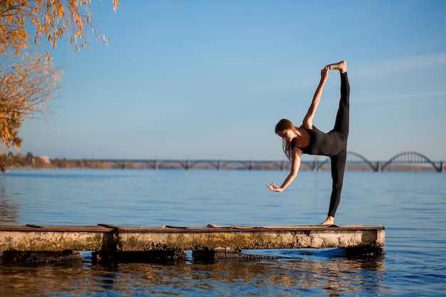 Mujer joven practicando ejercicio de yoga en el tranquilo muelle de madera con fondo de ciudad. Deporte y recreación en la ciudad