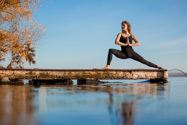 Mujer joven practicando ejercicio de yoga en el tranquilo muelle de madera con fondo de ciudad. Deporte y recreación en la ciudad