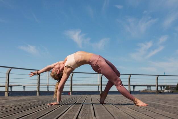 Mujer joven practicando la postura de yoga cosa salvaje en la terraza del paseo marítimo de madera en un día soleado
