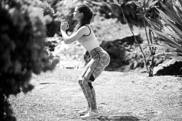 Mujer joven practicando sentadillas en el parque chica en forma haciendo ejercicio al aire libre en el día de verano hembra en forma trasera y