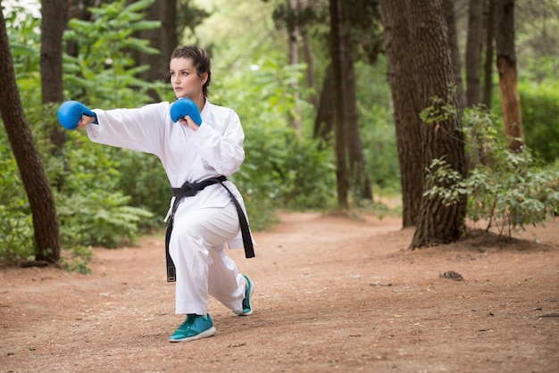 Mujer joven practicando sus movimientos de karate en el área del bosque arbolado Kimono blanco cinturón negro