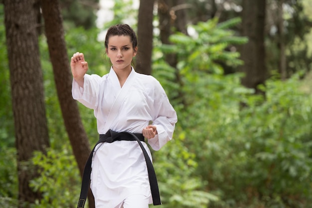 Mujer joven practicando sus movimientos de karate en el área del bosque arbolado Kimono blanco cinturón negro