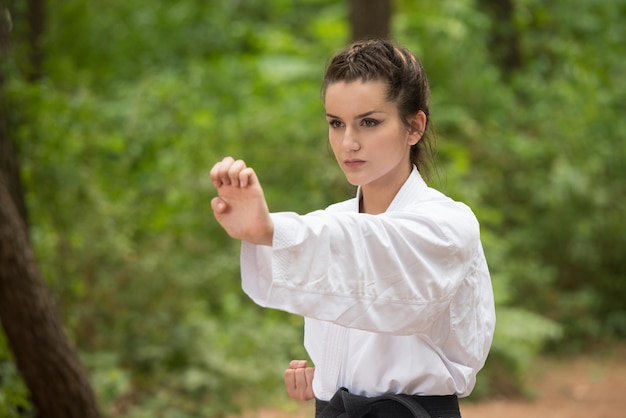 Mujer joven practicando sus movimientos de karate en el área del bosque arbolado Kimono blanco cinturón negro