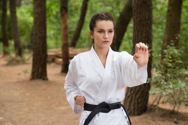 Mujer joven practicando sus movimientos de karate en el área del bosque arbolado Kimono blanco cinturón negro