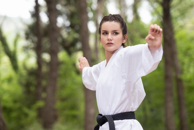 Mujer joven practicando sus movimientos de karate en el área del bosque arbolado Kimono blanco cinturón negro