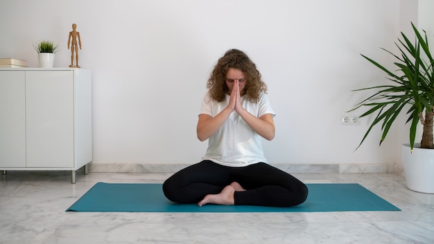 Mujer joven practicando yoga y haciendo el saludo Namaste sobre una alfombra azul en casa