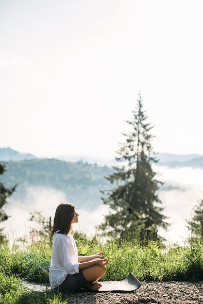 Mujer joven practicando yoga y sentada entre la hierba a la luz del sol en el fondo de las montañas de la mañana Meditación Tranquilo momento de conexión con la naturalezaZen Espacio de copia