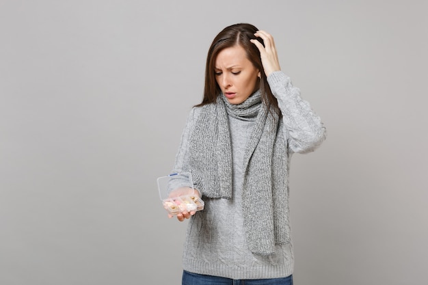 Mujer joven preocupada en suéter gris, bufanda puso la mano en la cabeza, sosteniendo mirando el pastillero diario aislado sobre fondo de pared gris. Estilo de vida saludable, tratamiento de enfermedades enfermas, concepto de estación fría.