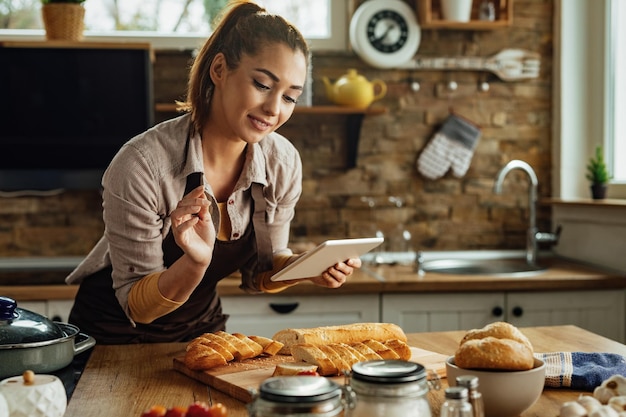 Mujer joven preparando comida mientras usa el panel táctil y sigue la receta en Internet en la cocina.