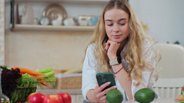 Mujer joven preparando ensalada buscando recetas en línea en el teléfono inteligente Preparando una ensalada vegana fresca y saludable Aplicaciones de menú de libros de cocina móviles Gente Tecnología moderna Concepto de cocina