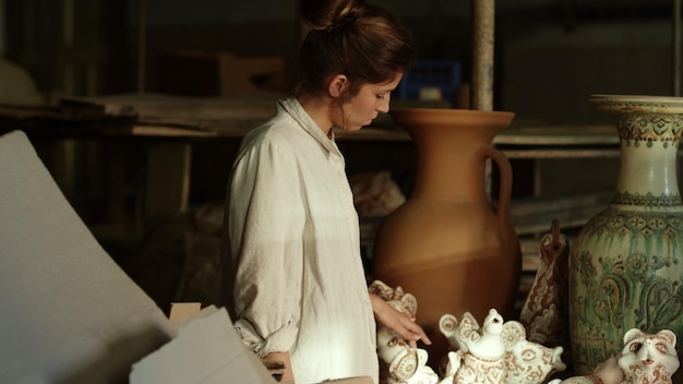 Mujer joven de primer plano examinando productos en cerámica Artista femenina seria tocando figuras de arcilla en el taller en cámara lenta Dama enfocada caminando en un estudio lleno de cerámica