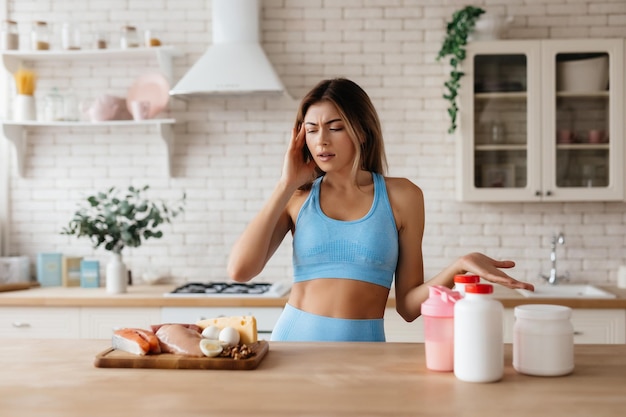 Mujer joven con problemas frunciendo el ceño mientras mira la selección de productos en la mesa y señala suplementos alimenticios