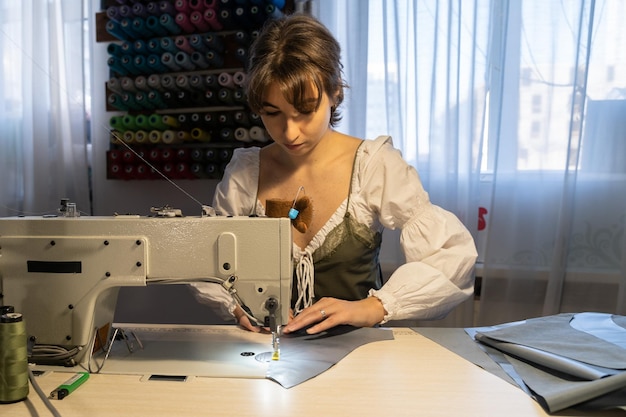 Mujer joven propietaria de un estudio de diseño en el lugar de trabajo cose ropa en una máquina de coser en un taller profesional