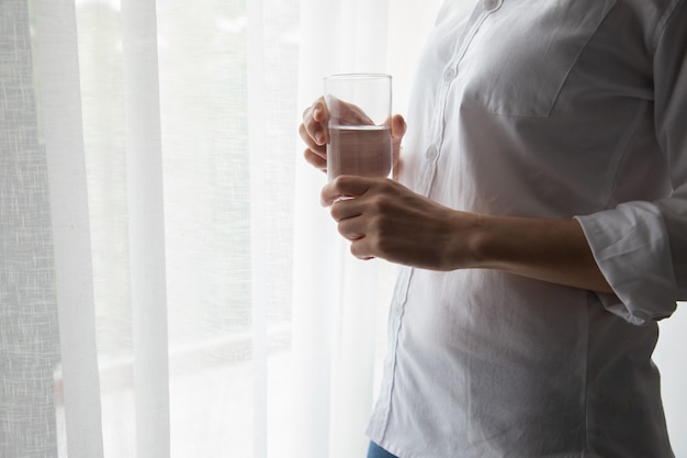 La mujer joven que bebe el agua mineral en las ventanas blancas de la cortina texturiza el fondo.