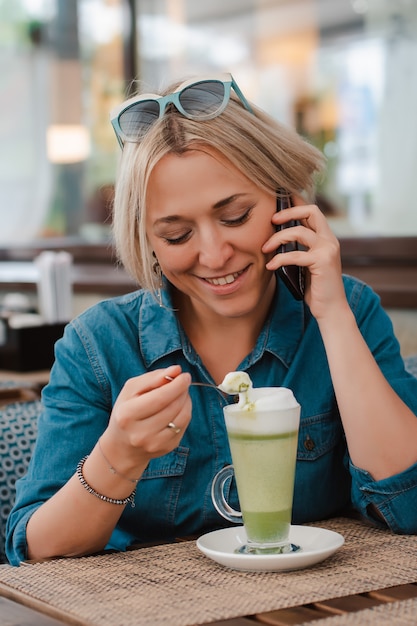 Mujer joven que bebe disfrutando del latte del té verde del matcha en mañana del verano en café.