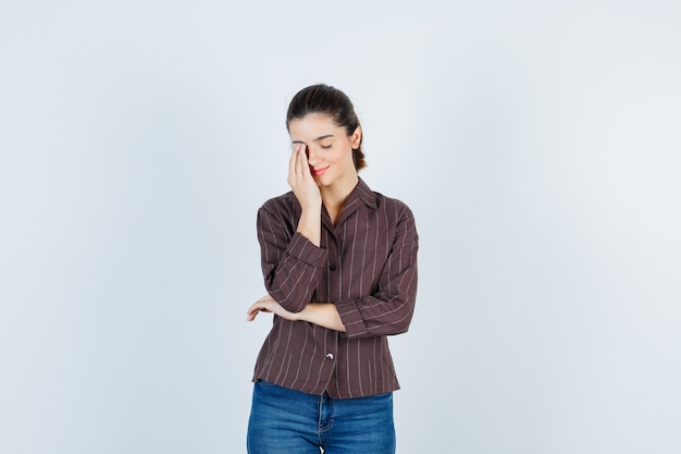 Mujer joven que cubre el ojo con la mano en camisa a rayas, jeans y mirando exhausto, vista frontal.