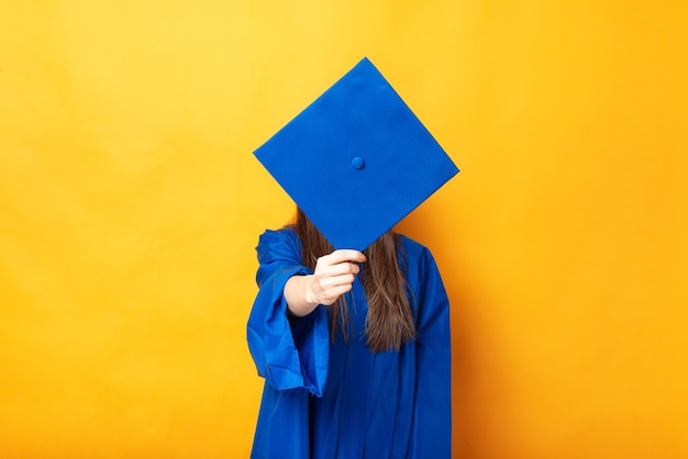 Foto mujer joven que cubre su rostro con gorro de graduación sobre fondo amarillo