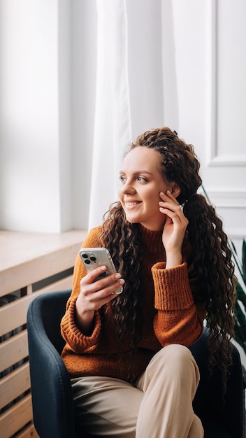 Mujer joven que disfruta de aplicaciones de teléfonos inteligentes y mensajes de texto mientras está sentada en una silla mujer usuaria de teléfono móvil