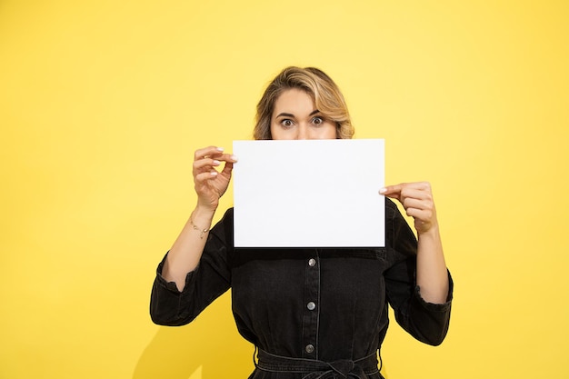 Mujer joven que se esconde detrás de la hoja de papel en blanco para la inscripción. Foto sobre fondo amarillo en estudio. Persona irreconocible.