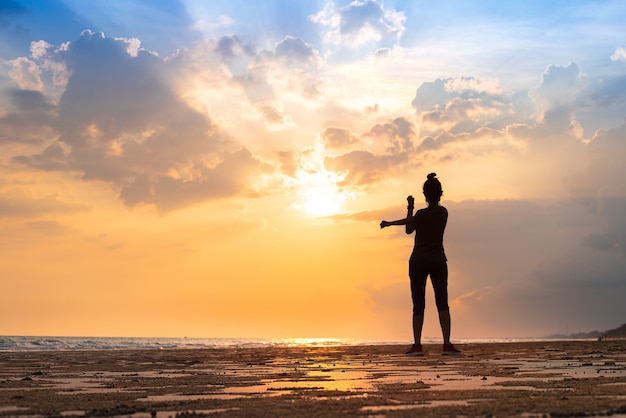 Mujer joven que se extiende para relajar su cuerpo después de correr, trotar y ejercitar la actividad para una buena salud en la playa y el mar en la hermosa puesta de sol y el cielo en la noche