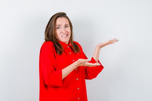 Foto mujer joven que finge mostrar algo en blusa roja y parece confundida. vista frontal.