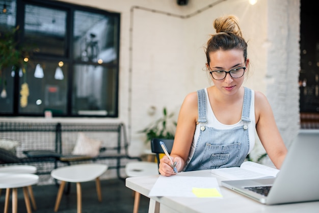 Mujer joven que hace notas y que estudia con el ordenador portátil en lugar público.