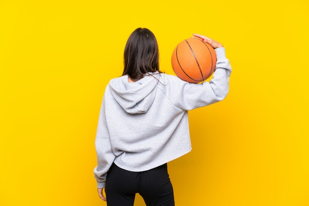 Mujer joven que juega a baloncesto sobre la pared amarilla aislada