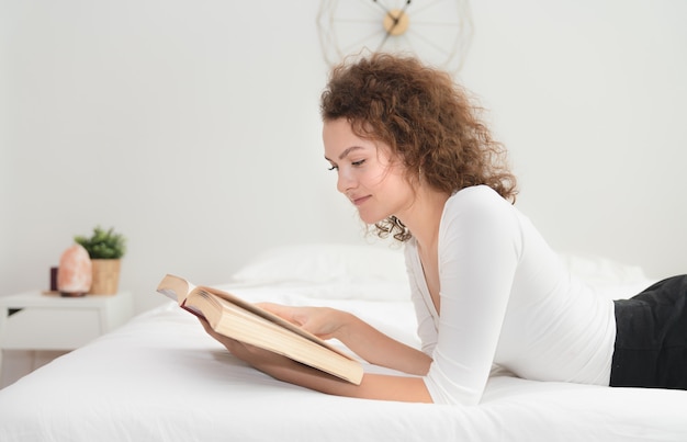 Mujer joven que lee un libro en la cama por la mañana