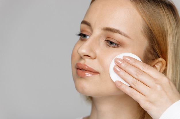 Mujer joven que limpia (quitando maquillaje) su cara con algodón, fondo gris. Cuidado de la piel saludable
