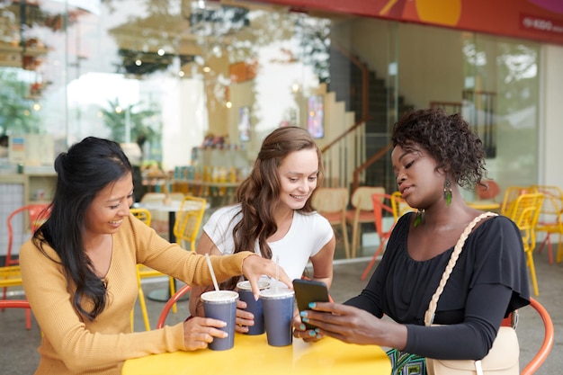 Mujer joven que muestra fotos en el teléfono inteligente a amigos cuando toman refrescos en la mesa del café