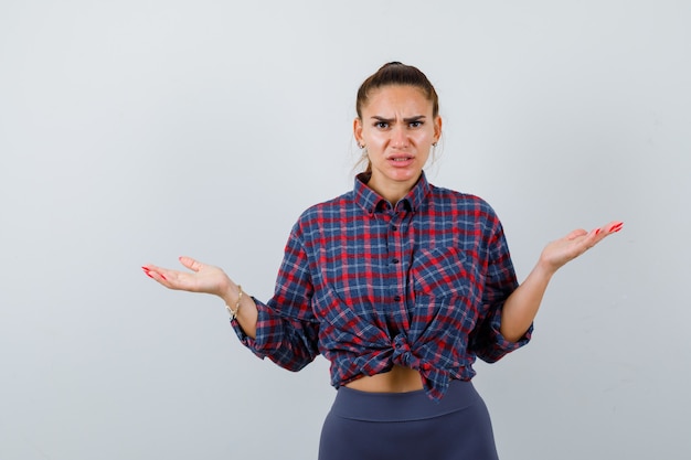 Mujer joven que muestra un gesto de impotencia en camisa a cuadros, pantalones y mirando estresado, vista frontal.