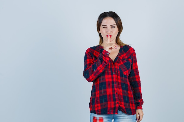 Mujer joven que muestra un gesto de silencio mientras parpadea en camisa a cuadros y mira confiada, vista frontal.