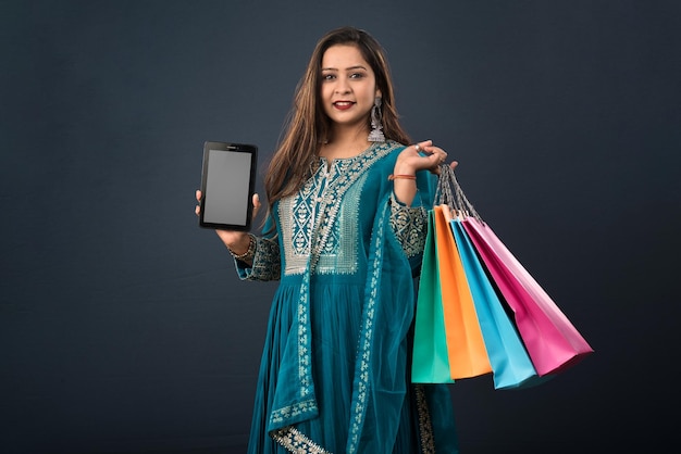 Mujer joven que muestra la pantalla en blanco del teléfono móvil o tableta y bolsas de compras en sus manos sobre un fondo gris