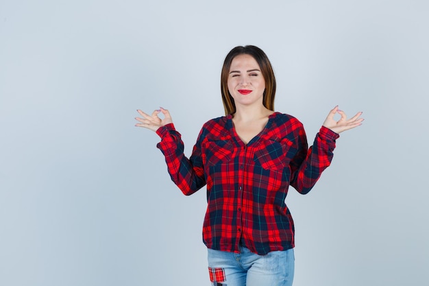 Mujer joven que muestra el signo de mudra mientras parpadea en camisa, jeans y mira positiva, vista frontal.