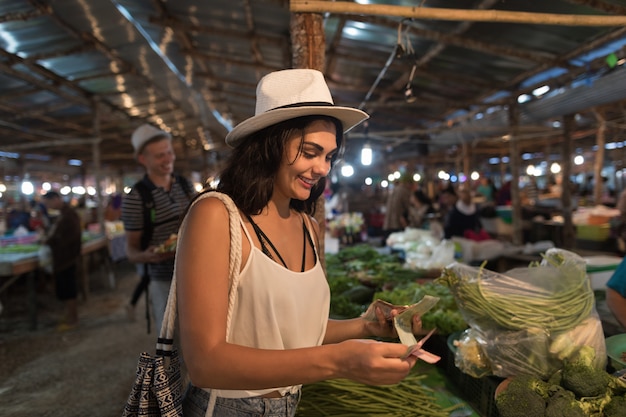 Foto mujer joven que paga dinero por las verduras frescas en el mercado chica de compras en el bazar de la calle