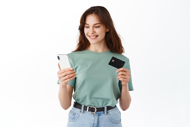Mujer joven que paga en línea con tarjeta de crédito escribiendo en un teléfono inteligente haciendo pedidos de compras en una aplicación móvil de pie contra el fondo blanco