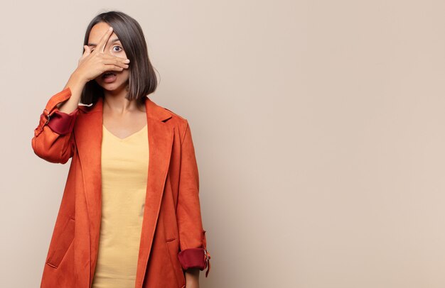 Mujer joven que parece sorprendida, asustada o aterrorizada, cubriendo la cara con la mano y mirando entre los dedos