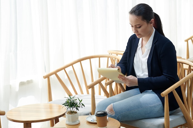 Mujer joven que pasa la mañana en la cafetería, tomando café y escribiendo pensamientos e ideas sobre el desarrollo empresarial en el cuaderno