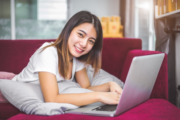 Mujer joven que practica surf el Internet por la computadora portátil en el sofá rojo en emoción alegre del humor del gesto