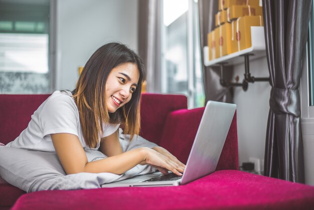 Mujer joven que practica surf el Internet por la computadora portátil en el sofá rojo en emoción alegre del humor del gesto