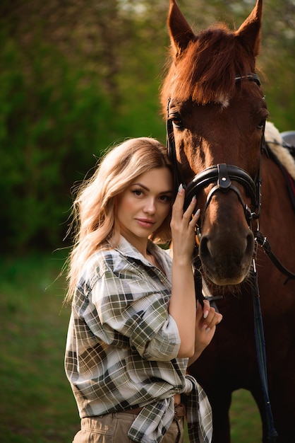 Mujer joven que presenta con un caballo marrón en un bosque en un prado soleado.