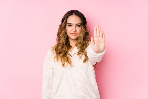 Mujer joven que presenta en una pared rosada que se coloca con la mano extendida que muestra la señal de stop, previniéndole.