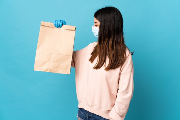 Mujer joven que protege del coronavirus con una máscara y sosteniendo una bolsa de compras aislada en azul con expresión feliz