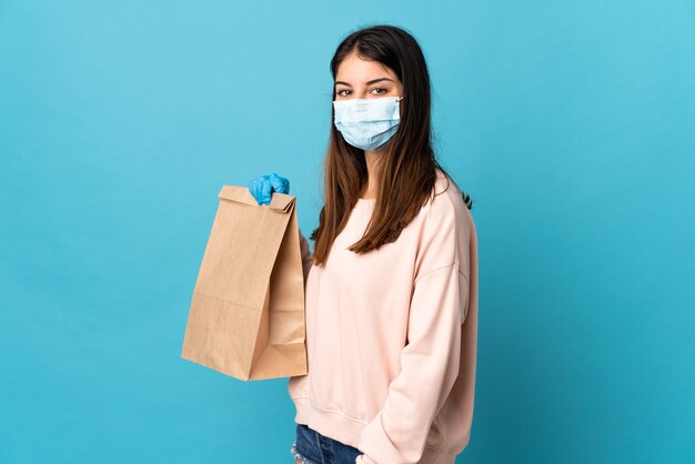 Mujer joven que se protege del coronavirus con una máscara y sostiene una bolsa de la compra aislada en azul sonriendo mucho