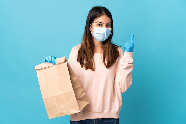 Mujer joven que se protege del coronavirus con una máscara y sostiene una bolsa de compras aislada en una pared azul que apunta hacia una gran idea